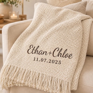 Op de afbeelding: Een crèmekleurige gebreide plaid met een franjerand en donkerbruine geborduurde tekst die Ethan + Chloe leest, gevolgd door de datum 11.07.2025. De deken heeft een getextureerd, geweven uiterlijk en is over een meubelstuk gedrapeerd.