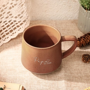 Pode incluir: Uma caneca de cerâmica com acabamento em terracota salpicado e uma alça lateral. A parte frontal da caneca apresenta o texto impresso em branco "Papa EST 2026" numa combinação de fonte manuscrita e sem serifa. Base larga e arredondada.