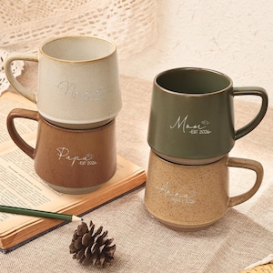 Può includere: Quattro tazze in ceramica impilate a coppie. Una coppia presenta una tazza beige con "Mom" e una tazza marrone con "Papa". L'altra coppia include una tazza verde con "Mom" e una tazza beige macchiata con "Mama". Tutte le tazze hanno manici.