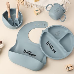May include: A baby feeding set in a soft blue color, including a bowl with a spoon and fork, a divided plate, a bib, and a sippy cup. The name "William" is printed on the plate and bib. Wooden blocks and a small succulent are also present.