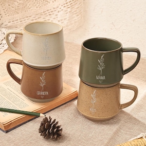 May include: Four ceramic mugs stacked in pairs. The top mug is beige with the word "Sophia" and a floral design. The bottom mug is brown with the word "Grandpa". The other pair includes a green mug with "Mama" and a tan mug with "Kelly".