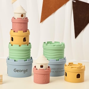 Puede incluir: Juego de juguetes de castillo apilables en colores pastel. El conjunto incluye una torre con techo blanco, secciones rosas, amarillas, verdes y azules. La base azul tiene el nombre "George" impreso. También se ven piezas de castillo adicionales.