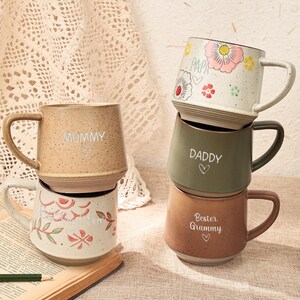 May include: A collection of ceramic mugs in shades of beige, green, and brown. Each mug has a handle and is inscribed with words like "Mummy," "Daddy," and "Best Granny." Some mugs are adorned with floral patterns and hearts.
