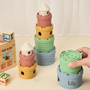 以下が含まれることがあります： パステルカラーの積み重ねタワーのおもちゃ。おもちゃは、ピンク、黄色、緑、青の色合いの城の形をしたいくつかの部分から構成されています。タワーの高さは約47.5cmです。木製のブロックセットも表示されています。