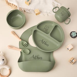May include: Sage green baby feeding set with a divided plate, bowl, bib, spoon, and fork. The set includes a sippy cup with handles. The plate and cup feature a bear graphic. The name "Arthur" is printed on the plate and bib.
