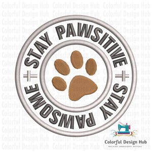 May include: Embroidered patch with a circular design. The text reads "STAY PAWSITIVE + STAY AWESOME". A brown paw print is in the center. The design is in shades of gray and brown.