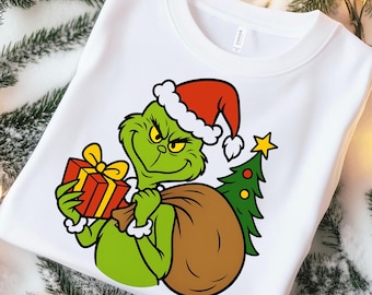May include: White sweatshirt featuring a green cartoon character wearing a Santa hat, holding a wrapped gift and a sack. A small Christmas tree with a star is also visible. The design is in the center of the shirt.