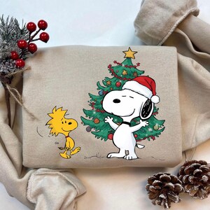 May include: Beige sweatshirt featuring a festive Snoopy design. Snoopy, wearing a Santa hat, stands in front of a decorated Christmas tree. Woodstock is also featured. The design is in bright colors, perfect for the holiday season.