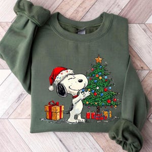 May include: Olive green sweatshirt featuring Snoopy wearing a Santa hat, hugging a decorated Christmas tree. Presents are at the base of the tree and to the left. The sweatshirt has long sleeves.