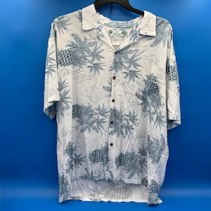 Può includere: Camicia bianca con bottoni con un motivo di ananas e foglie blu. La camicia ha maniche corte e un colletto. La camicia è appesa a una gruccia nera su sfondo blu. La camicia ha un'etichetta "Two Palms".
