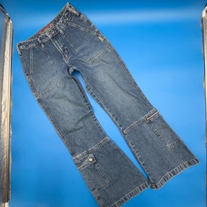 May include: Blue denim jeans featuring a high-waisted fit, two large front patch pockets, and a horizontal seam detail above the knee. The lower legs include a small zippered pocket on the left and a flap pocket on the right, with a flared silhouette.