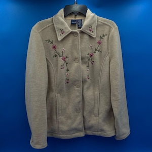 Puede incluir: Chaqueta de forro polar beige con cuello y cierre de botones. La chaqueta presenta bordados florales rosas y verdes. Tiene dos bolsillos delanteros y está colgada de una percha negra sobre un fondo azul.