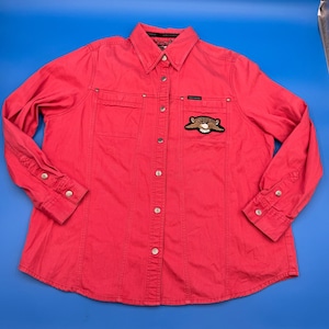 Może przedstawiać: Czerwona koszula zapinana na guziki z długimi rękawami. Koszula ma dwie kieszenie na piersi i naszywkę z motywem orła. Koszula ma srebrne guziki i kołnierzyk. Koszula jest wykonana z wytrzymałego materiału.
