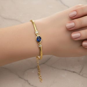 以下が含まれることがあります： 中央に青い楕円形の宝石が付いた、金色のブレスレット。ブレスレットは、テクスチャ加工された蛇のようなチェーンのデザインです。小さな透明な石が青い宝石の両側にセットされています。ブレスレットは手首に表示されています。