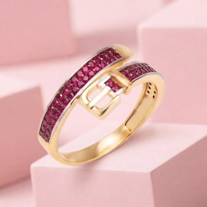 May include: A gold ring designed to resemble a belt, featuring a buckle-shaped centrepiece and a band encrusted with numerous small, vibrant red rubies. The metal has a polished finish, and the stones are set in a continuous row along the band.