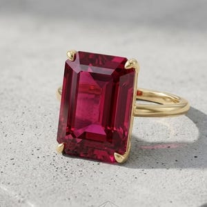 Op de afbeelding: Een gouden ring met een grote, rechthoekige, smaragdgeslepen dieproze edelsteen, gezet met vier pootjes. De ringband is van glad, gepolijst goud en houdt de levendige, gefacetteerde steen stevig op zijn plaats met een helder oppervlak.