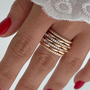 Könnte beinhalten: Ein goldener Ring-Set mit mehreren Bändern. Einige Bänder sind mit kleinen, klaren Edelsteinen besetzt, andere weisen ein gedrehtes Seil-Design auf. Die Hand trägt roten Nagellack.