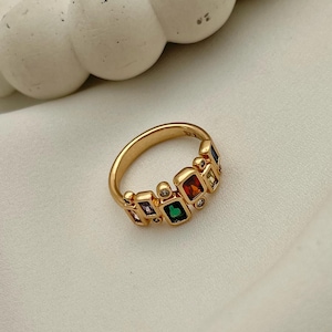 May include: A gold-colored ring with a series of colorful, square-cut gemstones. The gemstones are arranged in a row and include red, green, yellow, and purple stones. Small, clear stones are interspersed between the larger gems.