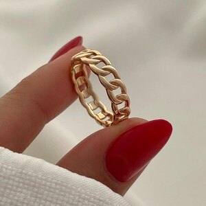 May include: A gold-colored chain link ring. The ring features a continuous chain design, with each link clearly defined. The ring is held against a white background, highlighting its metallic sheen. The ring is a piece of jewelry.