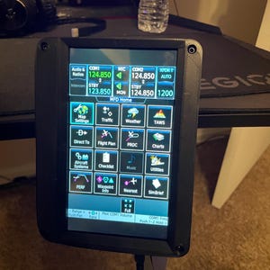 May include: A black electronic device with a screen displaying aviation information. The screen shows various options, including radio frequencies, map settings, and flight plan details. The device has a black frame and is connected to cables.