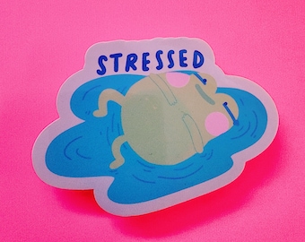 May include: A sticker featuring a cartoon frog floating in blue water, with the word "STRESSED" above. The frog is light green with pink cheeks and closed eyes. The sticker is on a bright pink background.