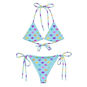 May include: A light blue bikini set with a heart pattern. The triangle top and tie-side bottoms feature colorful heart graphics with text. The straps and ties have a striped design in purple, yellow, and pink.