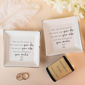 Könnte beinhalten: Zwei weiße quadratische Schmuckschalen mit goldenem Rand, jeweils mit einem motivierenden Zitat und einem Herzen versehen. Eine kleine Sojawachskerze und zwei goldene Ringe sind ebenfalls vorhanden. Die Kerze trägt die Aufschrift "ONLY FOR YOU".