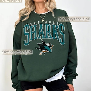 May include: A forest green crewneck sweatshirt featuring the text SAN JOSE SHARKS in a teal and white collegiate font, with a San Jose Sharks hockey logo graphic below the text. The sweatshirt is worn over a white shirt and black shorts.
