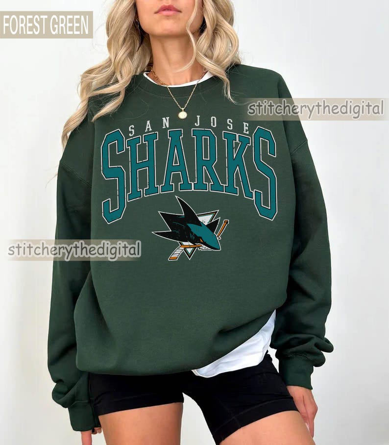 May include: A forest green crewneck sweatshirt featuring the text SAN JOSE SHARKS in a teal and white collegiate font, with a San Jose Sharks hockey logo graphic below the text. The sweatshirt is worn over a white shirt and black shorts.