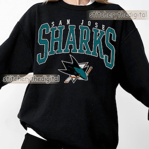May include: A black long-sleeve sweatshirt featuring the text SAN JOSE SHARKS in teal and white block lettering. Below the text is a graphic of a shark biting a hockey stick. The word BLACK is in the top left corner and stitcherythedigital is visible.
