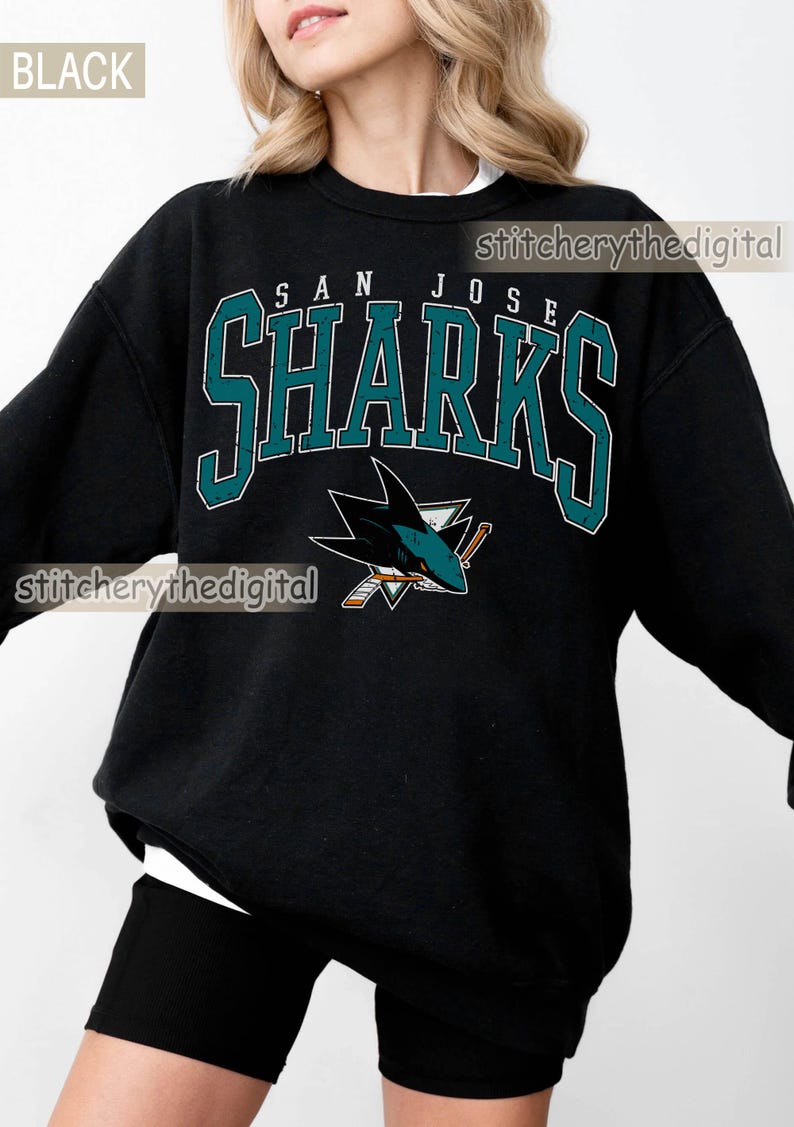 May include: A black long-sleeve sweatshirt featuring the text SAN JOSE SHARKS in teal and white block lettering. Below the text is a graphic of a shark biting a hockey stick. The word BLACK is in the top left corner and stitcherythedigital is visible.