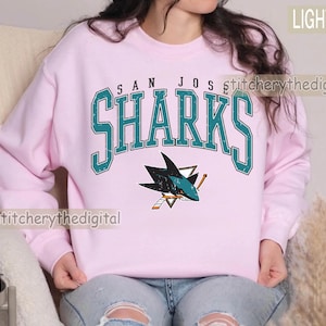 May include: A light pink crewneck sweatshirt featuring the text SAN JOSE SHARKS in a distressed teal font with a white outline. Below the text is a graphic of a shark biting a hockey stick, rendered in teal, black, white, and orange colors.