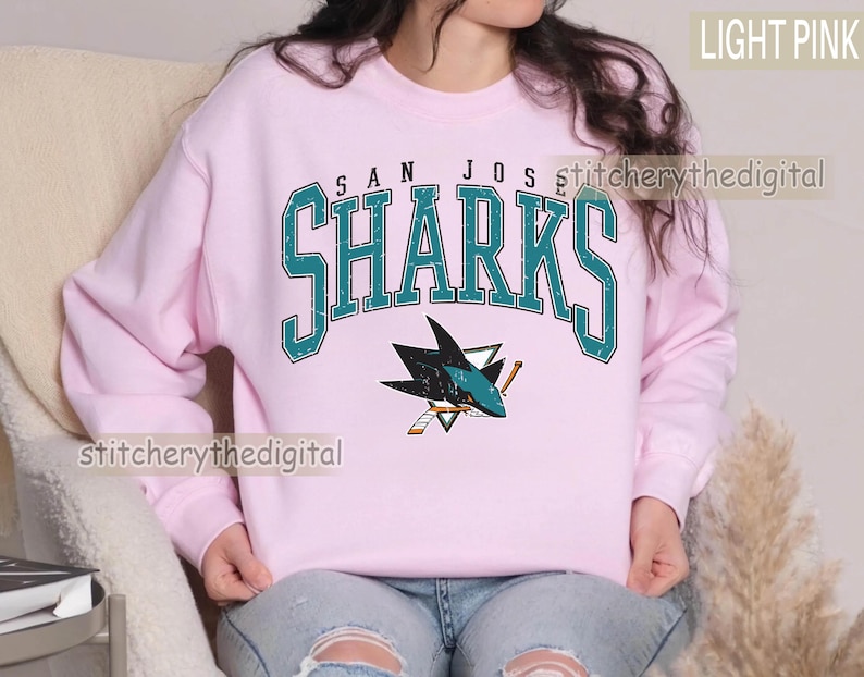 May include: A light pink crewneck sweatshirt featuring the text SAN JOSE SHARKS in a distressed teal font with a white outline. Below the text is a graphic of a shark biting a hockey stick, rendered in teal, black, white, and orange colors.