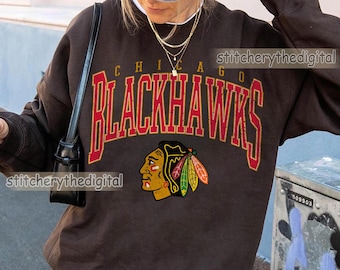 May include: A dark chocolate brown crewneck sweatshirt featuring the text CHICAGO BLACKHAWKS in a large red and yellow arched font. Below the text is a graphic of a Native American profile with colorful feathers in red, yellow, and green.