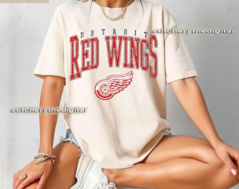 May include: An ivory short-sleeve t-shirt featuring the text DETROIT RED WINGS in a distressed red font across the chest. Below the text is a red graphic of a winged wheel logo. The shirt has a crew neckline and a relaxed, loose-fitting cut.