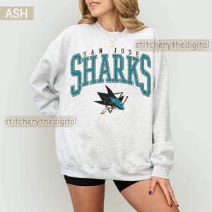 May include: An ash gray crewneck sweatshirt featuring the text SAN JOSE SHARKS in a distressed teal and black font. Below the text is a graphic of a shark biting a hockey stick. The sweatshirt has long sleeves and a ribbed collar and cuffs.