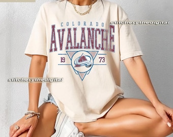 May include: Ivory-colored t-shirt featuring the text "COLORADO AVALANCHE" in a distressed, collegiate-style font. Below the text is a logo with a mountain and the numbers "19" and "73". The shirt has short sleeves and a relaxed fit.