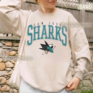 May include: A sand-colored crewneck sweatshirt featuring the text SAN JOSE SHARKS in a distressed teal font with a black outline. Below the text is a graphic of a shark biting a hockey stick, rendered in teal, black, and white colors.