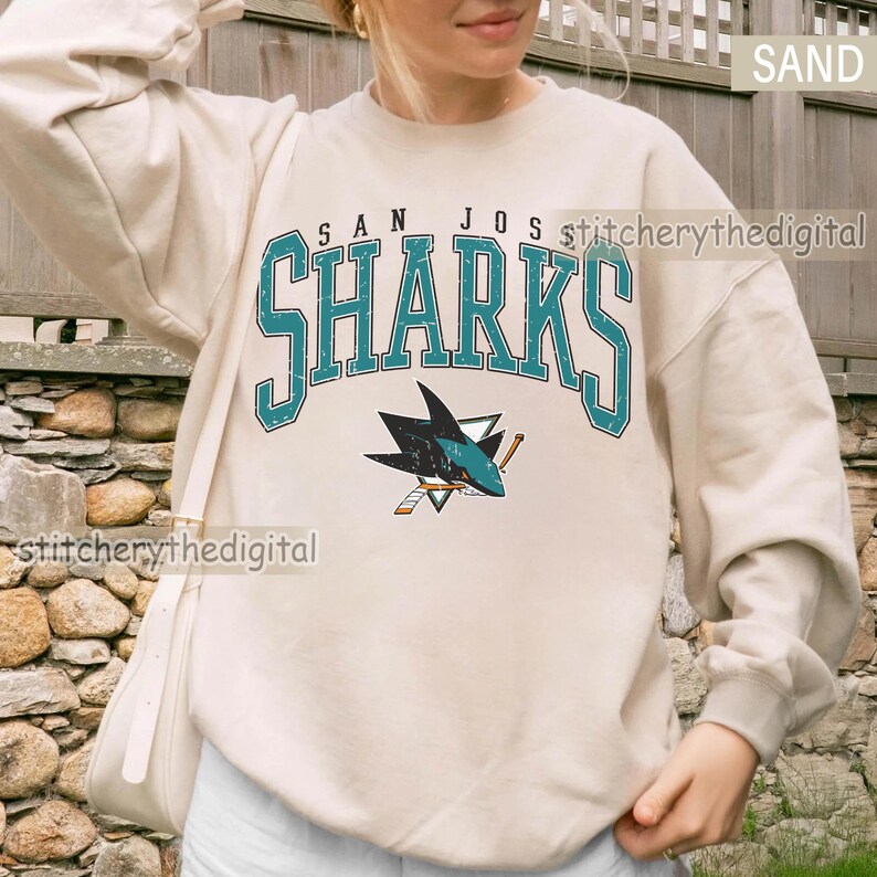May include: A sand-colored crewneck sweatshirt featuring the text SAN JOSE SHARKS in a distressed teal font with a black outline. Below the text is a graphic of a shark biting a hockey stick, rendered in teal, black, and white colors.
