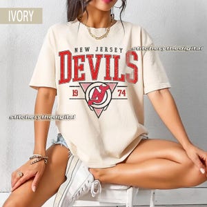 May include: Ivory-colored t-shirt featuring the text "NEW JERSEY DEVILS" in red, with a logo including a red "N" inside a circle and a triangle. The shirt also has the numbers "19" and "74" on either side of the logo. The shirt is short-sleeved.