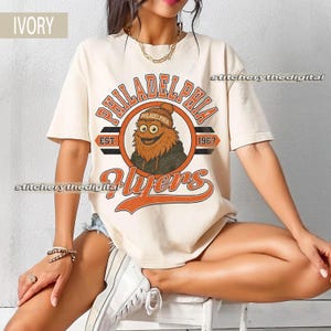 Può includere: T-shirt color avorio con un design dei Philadelphia Flyers. Il design include il nome della squadra, un'illustrazione della mascotte e l'anno di fondazione, 1967, in arancione e nero. La maglietta ha maniche corte e girocollo.