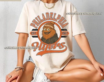 May include: Ivory t-shirt with a Philadelphia Flyers design. The design features the team name, a mascot illustration, and the year 1967 in orange and black. The shirt has short sleeves and a crew neck.