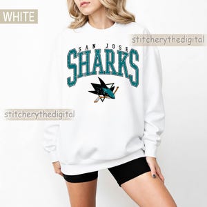 May include: A white long-sleeve crewneck sweatshirt featuring the text SAN JOSE SHARKS in a distressed teal and black font. Below the text is a graphic of a shark with a hockey stick. The sweatshirt has a relaxed fit and long sleeves.