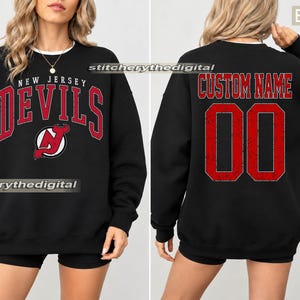 May include: A black crewneck sweatshirt featuring the New Jersey Devils logo and text on the front in red and white. The back displays the text CUSTOM NAME above the number 00 in a distressed red font with a white outline on the black fabric.