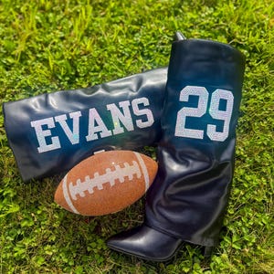 May include: Black leather boots with the number 29 in rhinestones, a matching clutch with the name EVANS in rhinestones, and a bedazzled football, all on a bed of green grass. The boots have a pointed toe and a mid-height heel.