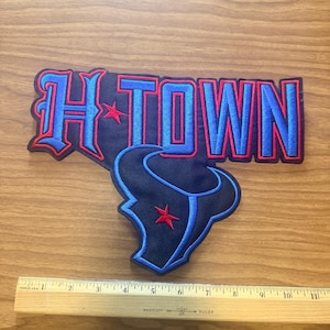 May include: A navy blue embroidered patch featuring the text H-TOWN in blue with red outlines and a red star, positioned above a navy blue bull head logo with a blue outline and a red star. A wooden ruler shows the patch is 11 inches wide.