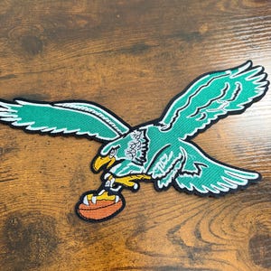 May include: An embroidered fabric patch featuring a green eagle in flight with white and black detailing. The eagle has a yellow beak and talons, and it is clutching an orange American football with white laces in its claws, outlined in black.