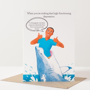 Peut inclure: Carte de vœux avec une illustration de dessin animé d'une personne faisant deux pouces levés à l'intérieur de la gueule d'un requin. La carte indique "Quand vous gérez cette dépression à haut fonctionnement". La bulle de dialogue dit "Je vais très bien ici, tout le monde, absolument bien. Comment va tout le monde ?"