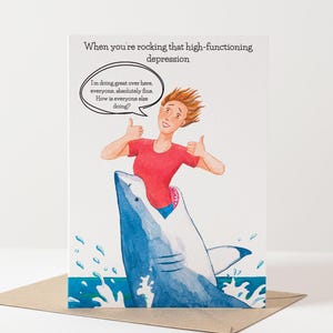 May include: Greeting card featuring a watercolour illustration of a person riding a blue shark, giving two thumbs up. The card reads "When you're rocking that high-functioning depression." A speech bubble says, "I'm doing great over here, everyone, absolutely fine. How is everyone else doing?"