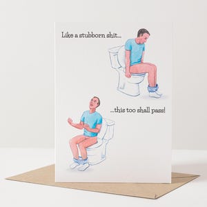 Può includere: Biglietto di auguri umoristico con un'illustrazione di una persona sulla toilette, con il testo "Like a stubborn shit... this too shall pass!" Il biglietto è su una busta marrone.